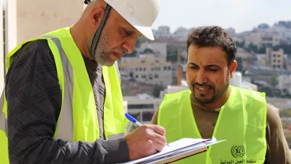 A man in a hard hat taking notes on a paper, while another watches with a smile, in Jordan